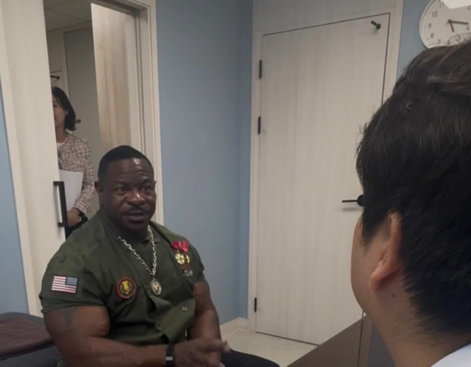 A patient seated in a clinic consultation room in Seoul, talking with a doctor whose back is to the camera, while a coordinator waits in the doorway.